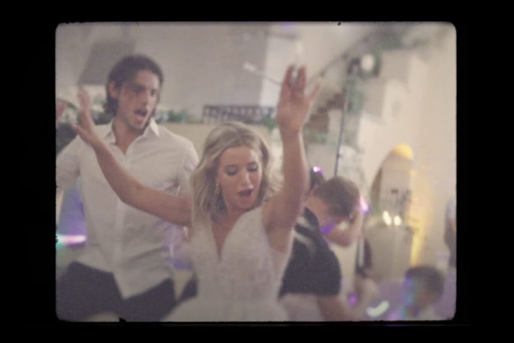 Wedding reception scene filmed with shallow depth of field, showing a bride dancing with raised arms and guests moving behind her, demonstrating videography definition through motion, lighting, and cinematic focus.