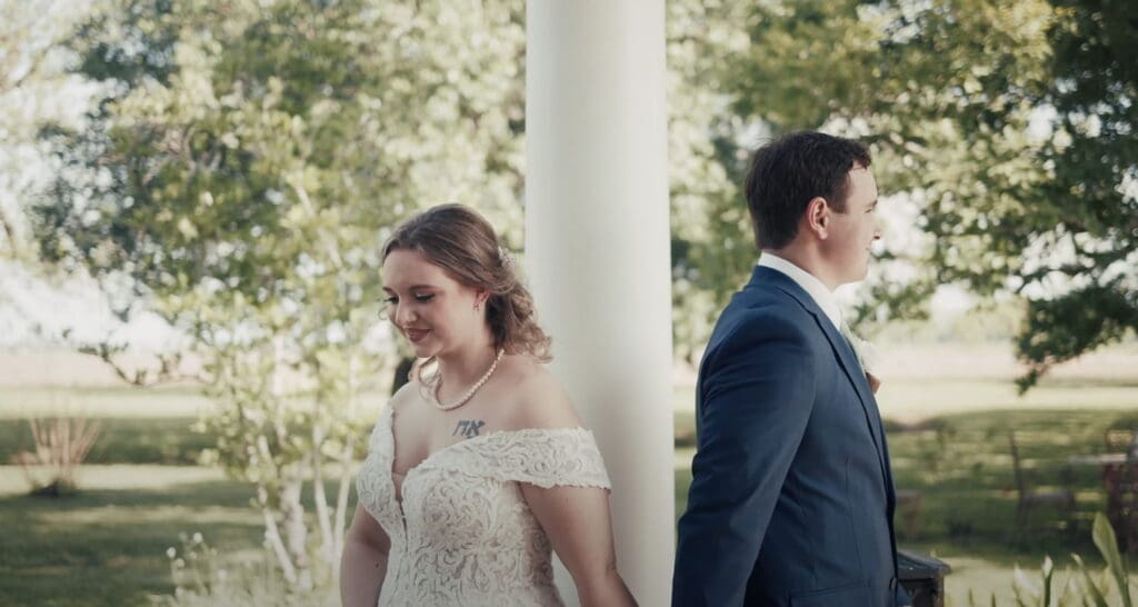 Bride and groom stand back to back on opposite sides of a white column outdoors before the ceremony, capturing an emotional first look moment included in wedding video packages.