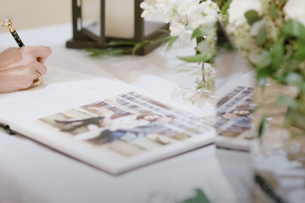 Guest signs a wedding book with a pen beside photos and floral decor, often included during wedding toasts
