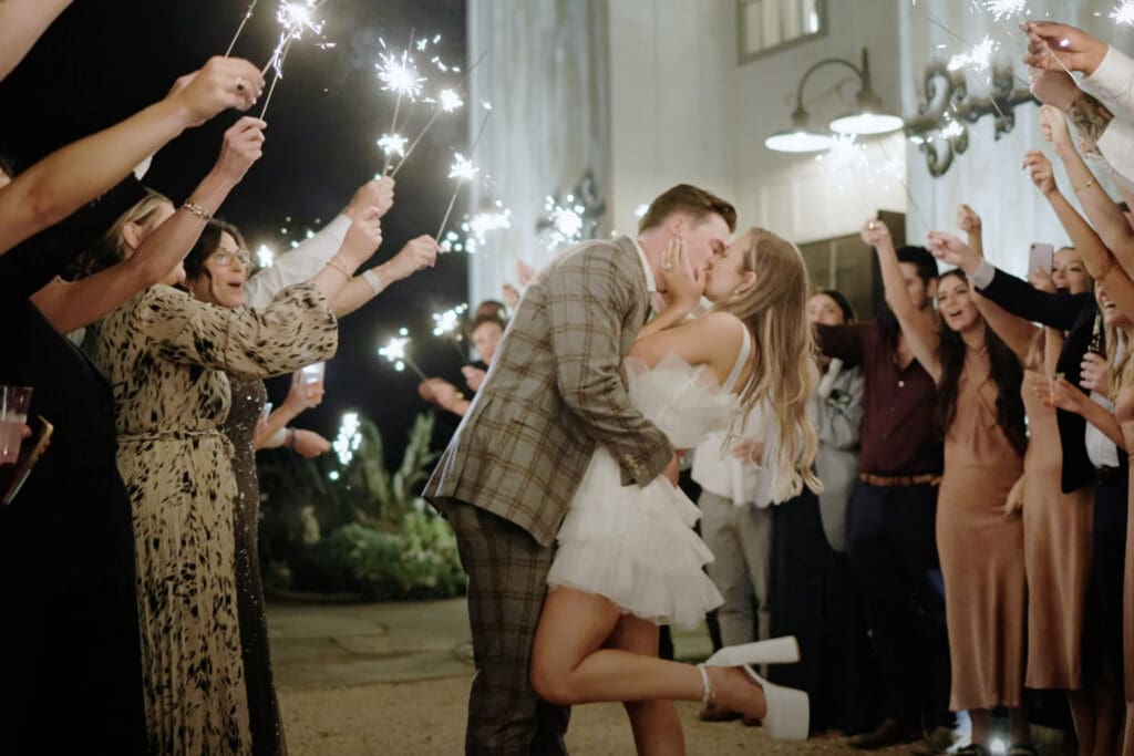 Bride and groom kiss while guests hold sparklers in formal attire, helpful visual for what to wear to a casual wedding