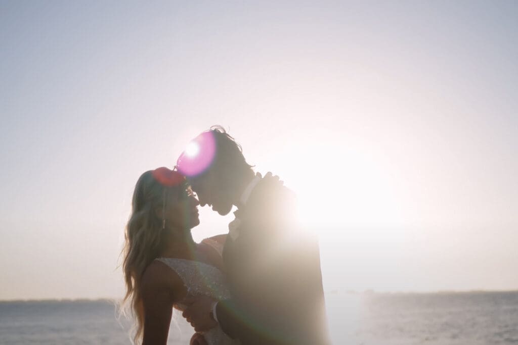 Romantic beach sunset engagement photo ideas showing a couple embracing in silhouette by the ocean with soft light and lens flare.