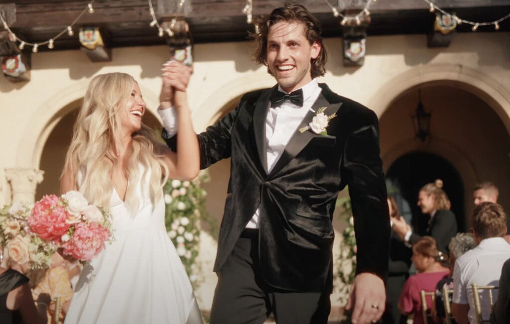 Bride and groom walk hand in hand down the aisle after the ceremony, smiling and celebrating with guests in the background. The bride wears a white gown and holds a pink bouquet, while the groom wears a black tuxedo with a boutonniere. Warm sunlight and string lights create a joyful, romantic setting that reflects thoughtful wedding planning and a well-organized celebration.