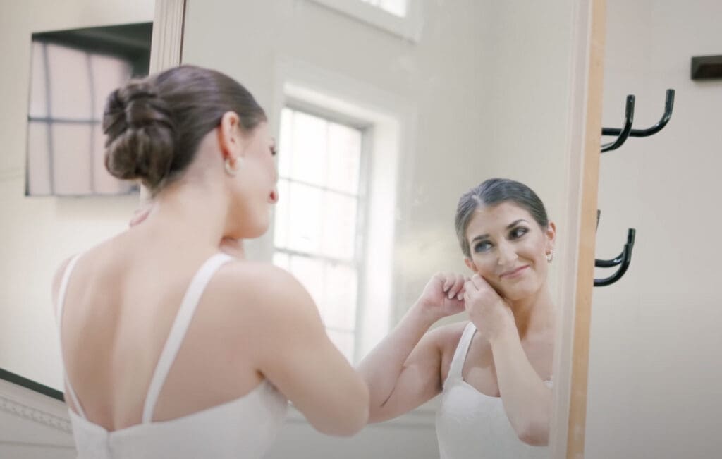 Bride stands in front of a mirror and adjusts her earring while wearing a white dress, with her hair styled in a neat bun. Soft natural light fills the room and highlights her calm, focused expression. This moment reflects a wedding planning checklist step as the bride completes final getting-ready details before the ceremony.