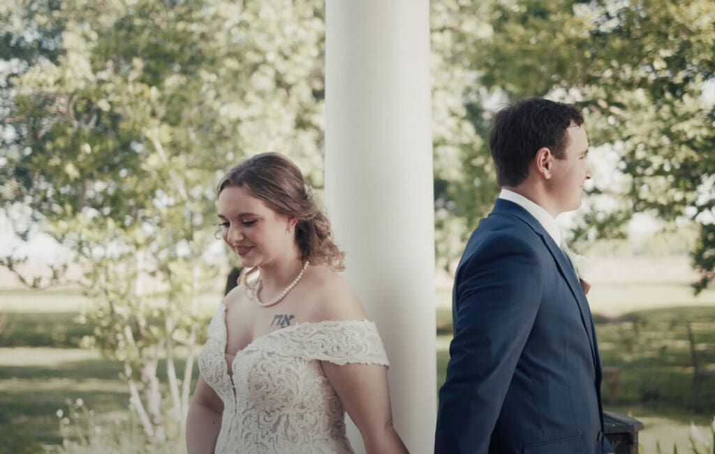 Bride and groom stand back to back outdoors before their first look, each facing opposite directions near a white column. The bride wears an off-shoulder lace gown and pearl necklace, while the groom wears a blue suit with a boutonniere. Trees and soft daylight create a calm setting that reflects a well-planned wedding planning timeline leading up to this emotional moment.
