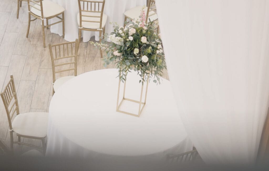 Round reception table with a white tablecloth and a tall floral centerpiece in a gold stand, surrounded by gold chiavari chairs in a bright indoor venue. Soft natural light and neutral decor create a clean, elegant setup that reflects careful wedding planning timetable coordination for reception design and layout.
