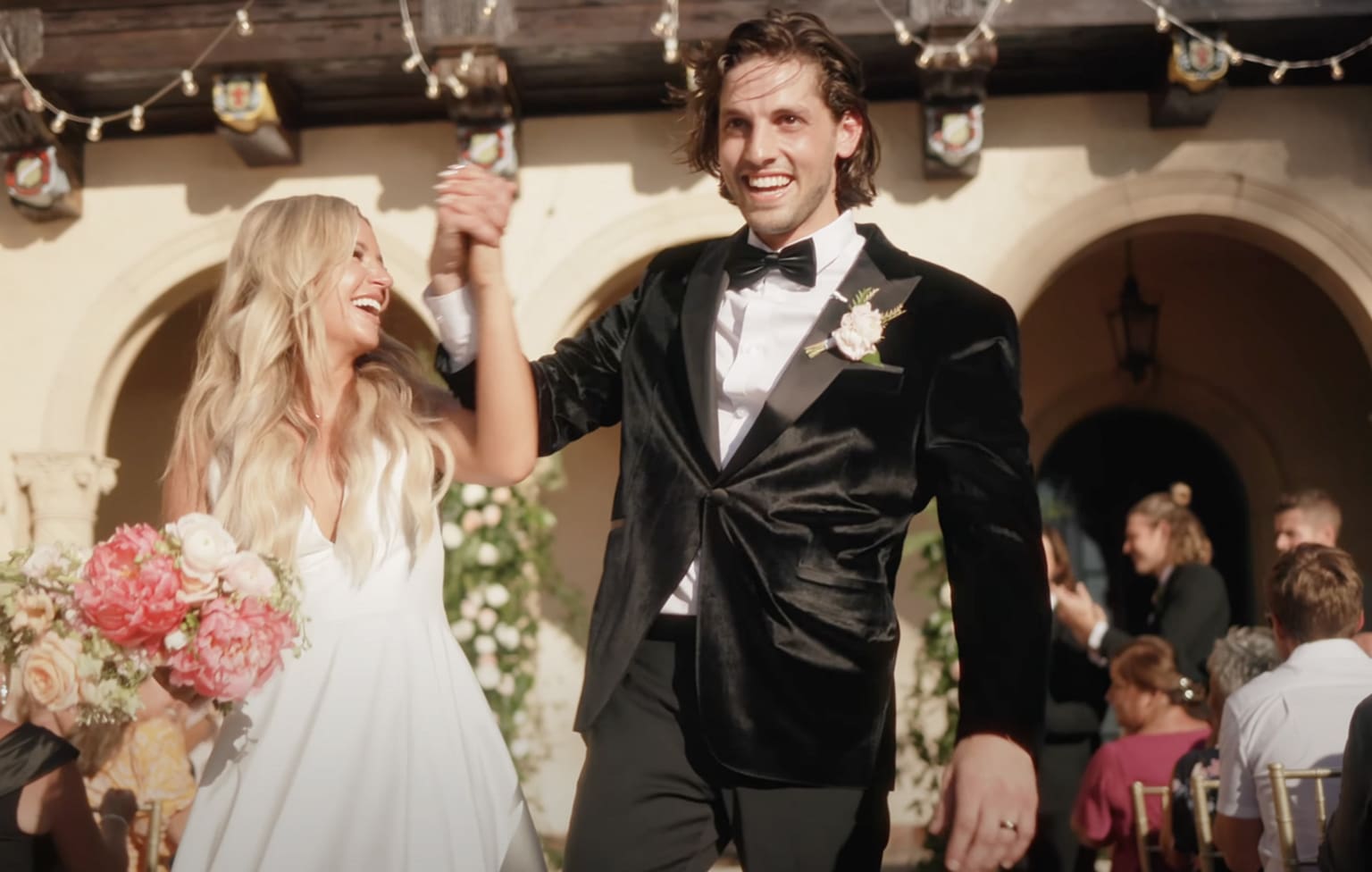 Bride and groom walk hand in hand down the aisle after the ceremony, smiling and celebrating with guests in the background. The bride wears a white gown and holds a pink bouquet, while the groom wears a black tuxedo with a boutonniere. Warm sunlight and string lights create a joyful, romantic setting that reflects thoughtful wedding planning and a well-organized celebration.