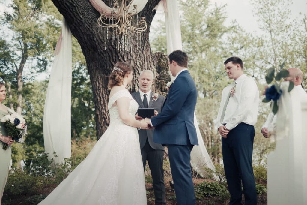 Outdoor wedding ceremony under a large tree where the bride and groom hold hands as the officiant speaks, with wedding party standing nearby, illustrating how to write wedding vows for a meaningful ceremony.