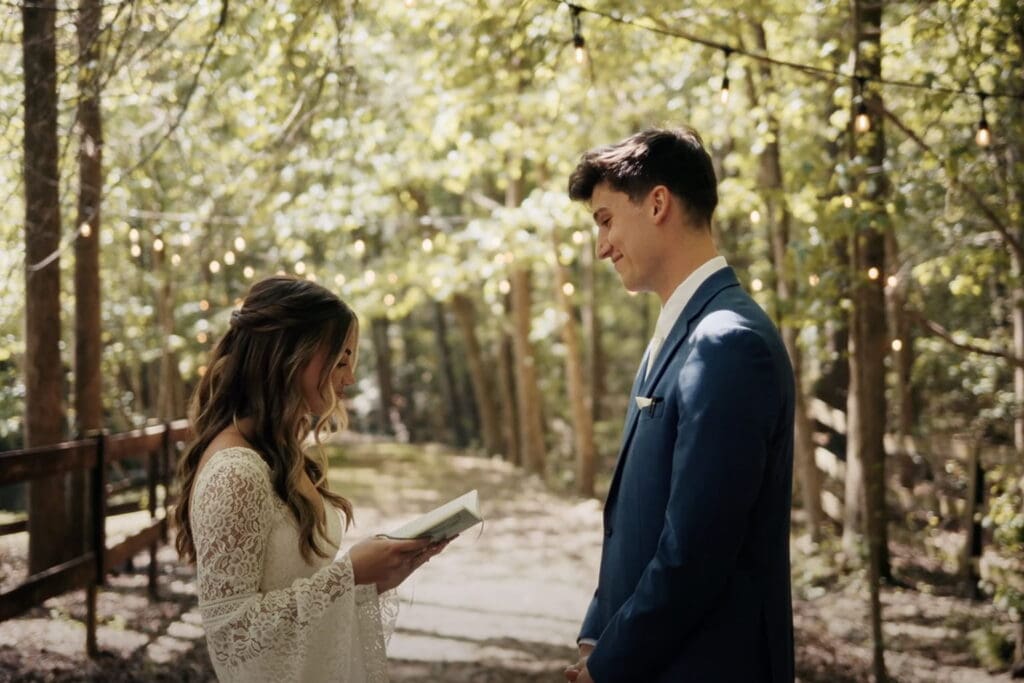 Bride reads wedding vows from a small book while facing the groom on a sunlit wooded path, with string lights hanging overhead and a rustic outdoor setting creating a warm, intimate ceremony atmosphere.