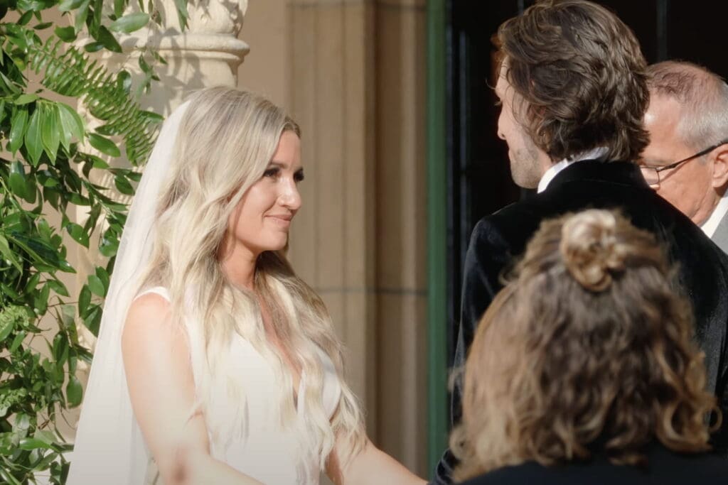 Bride and groom stand face to face during an outdoor ceremony beside a floral arch, smiling as the officiant prepares to speak, illustrating wedding vows examples in a romantic setting.