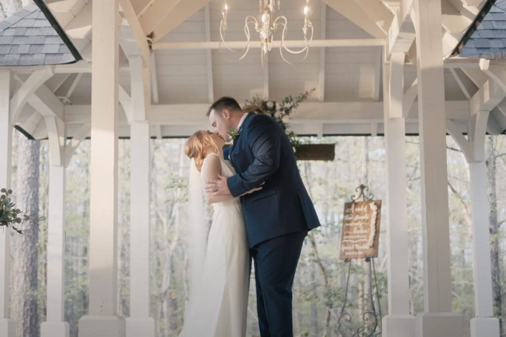 Bride and groom share a kiss under a white outdoor pavilion during their ceremony, with bridesmaids and a groomsman nearby, illustrating a romantic wedding vows script moment.